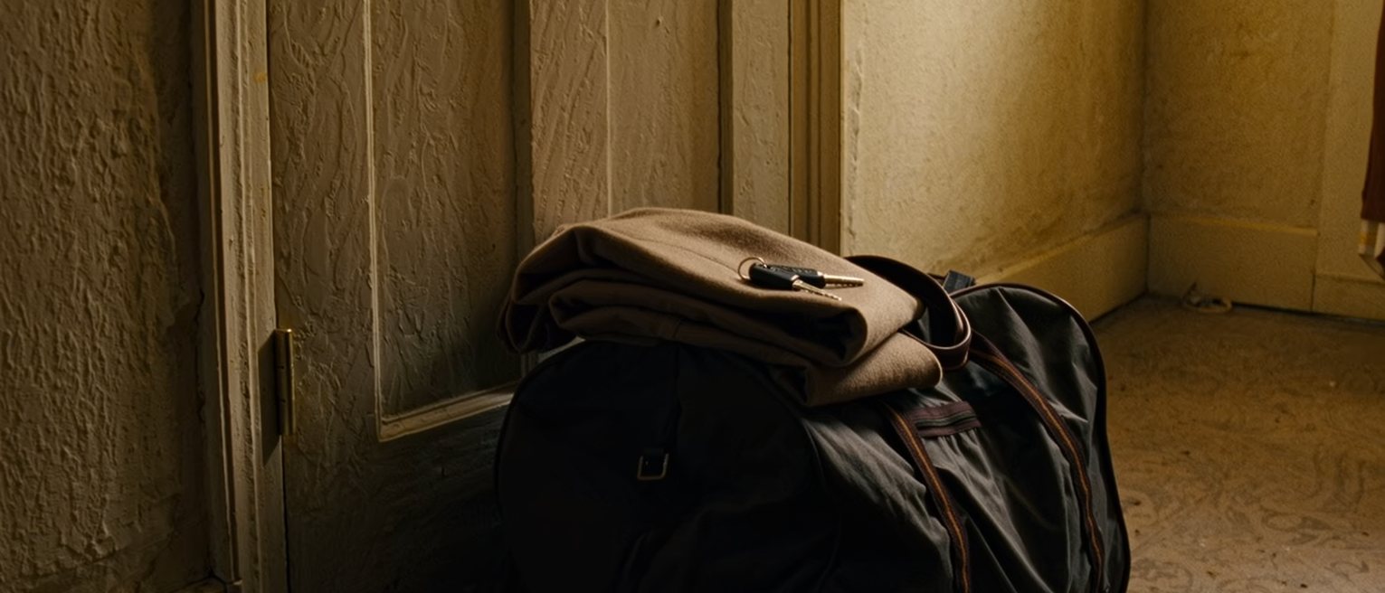 a hall seen close, a packed overnight bag set by the door with a coat folded on top of it and a set of keys laid on the bag, the door on the latch behind