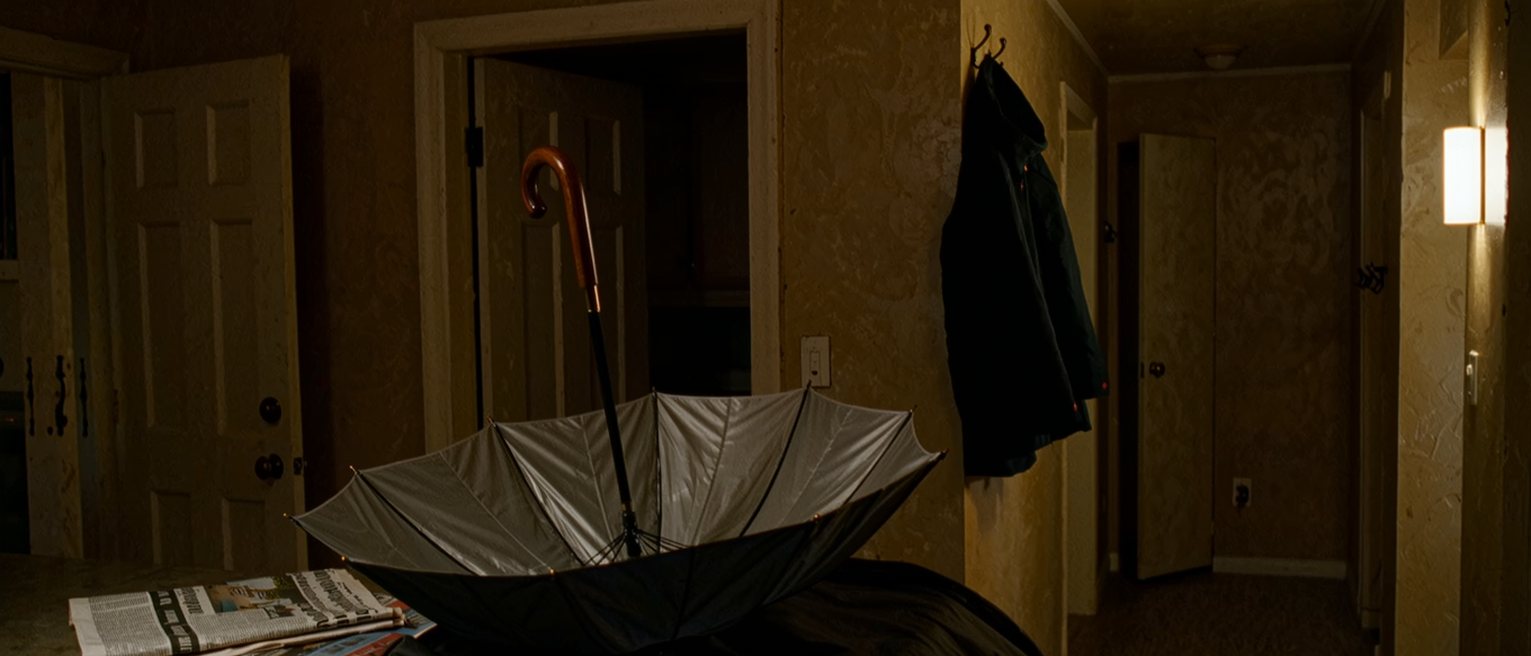 a kitchen counter with an umbrella left open to dry, a raincoat on the hook beside it and a folded weather page of the paper on the worktop, the window soft behind