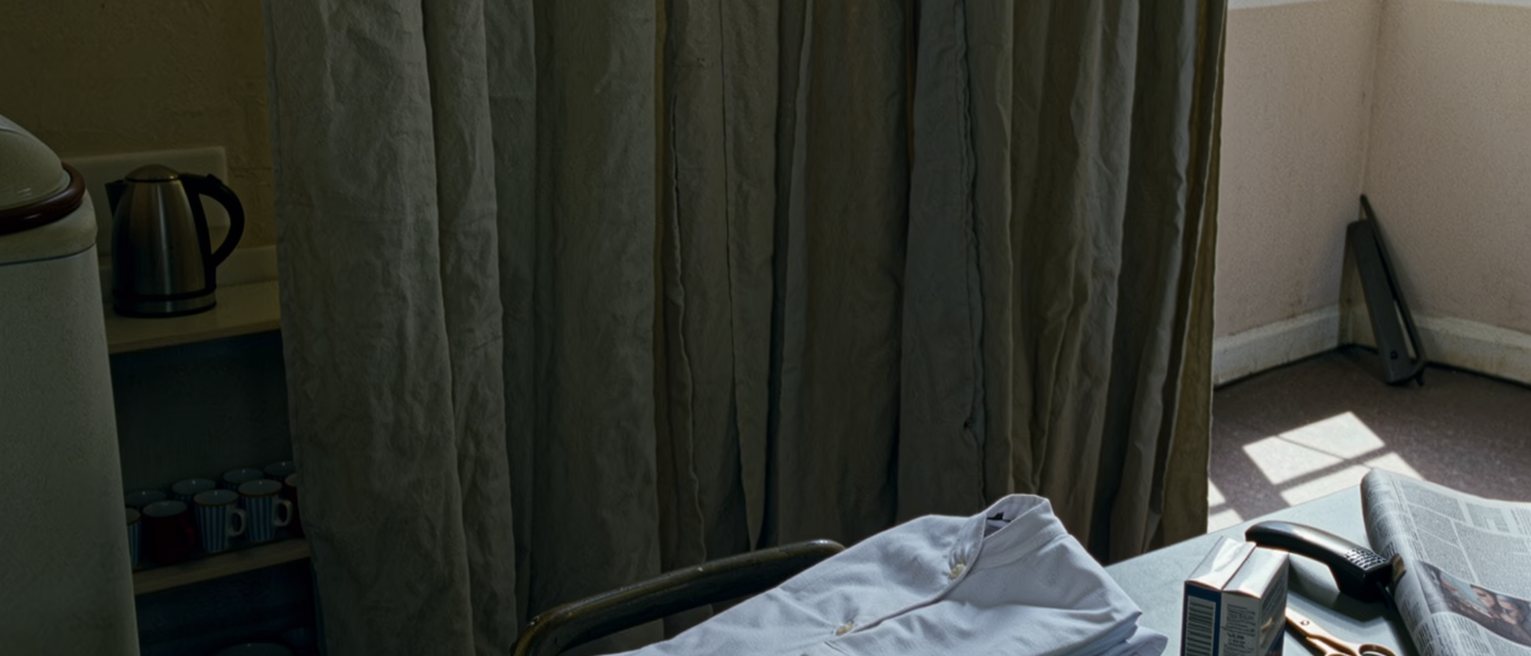 a bedsit seen close with a white uniform folded on the chair, a box of hair dye unopened beside a pair of scissors, a newspaper with a folded page and a phone face down, the kettle and the shelf of cups close behind and the curtains drawn beyond