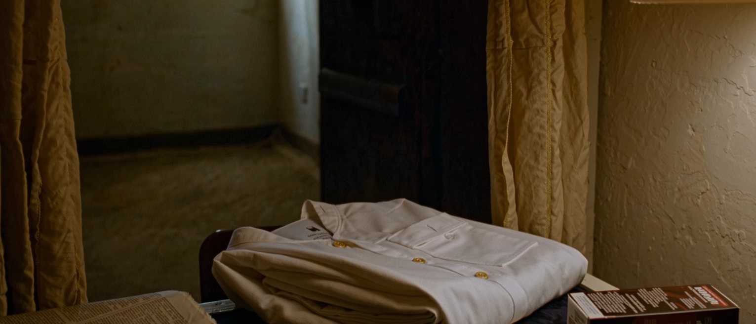 a bedsit seen close with a white uniform folded on the chair, a box of hair dye unopened beside a pair of scissors, a newspaper with a folded page and a phone face down, the kettle and the shelf of cups close behind and the curtains drawn beyond