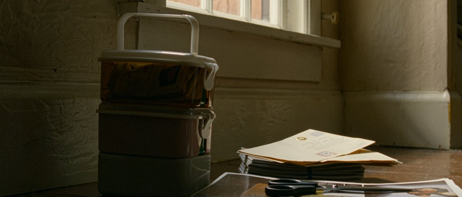 a kitchen seen close with a lunchbox packed for one, a stack of unopened letters and a pair of scissors beside a magazine with a page cut out, the window soft behind