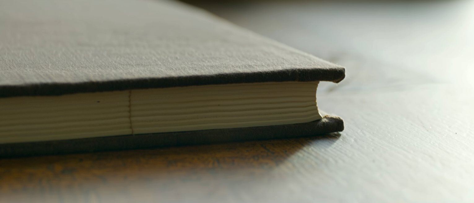 a thick book bound in dark cloth, closed and squared on a table, its page edges cut clean and even