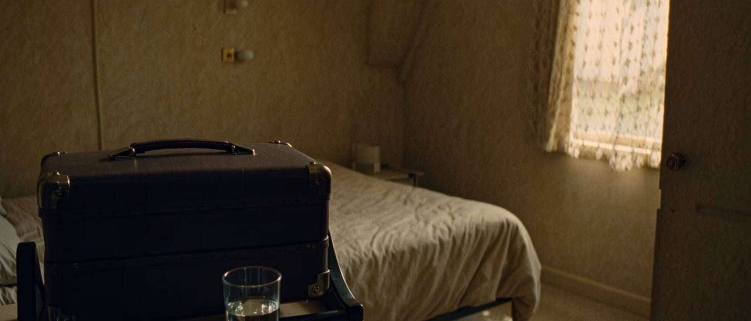 a bedroom seen close with a suitcase closed on the luggage stand, a room key on its fob and a folded receipt on the desk, a glass of water, the bed made tight close behind and the tall window with its net curtain soft beyond