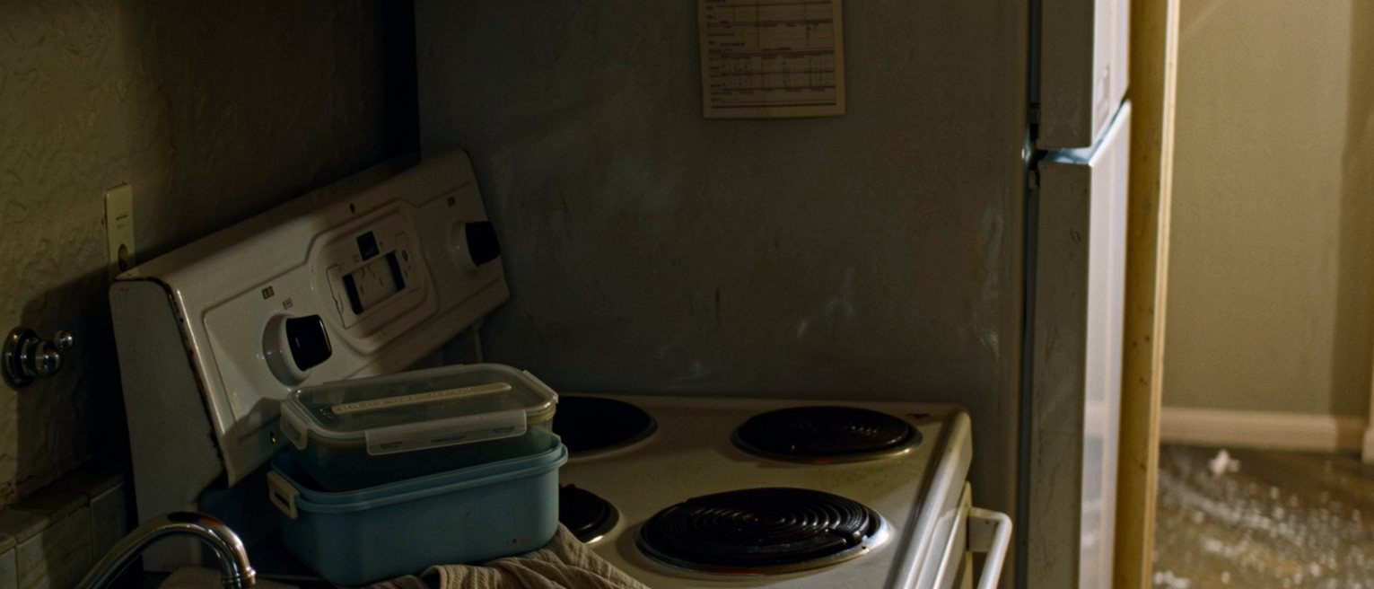 a kitchen seen close with a dishcloth folded over the tap, a lunch box washed and left to dry, a shopping list held on the fridge under a magnet, the stove close behind and the hallway soft beyond