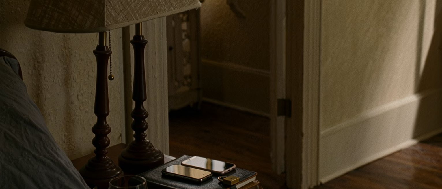 a bedside table seen close with two phones charging side by side, one face down and one face up, a diary with a small lock and a glass of water, the lamp and the headboard close behind