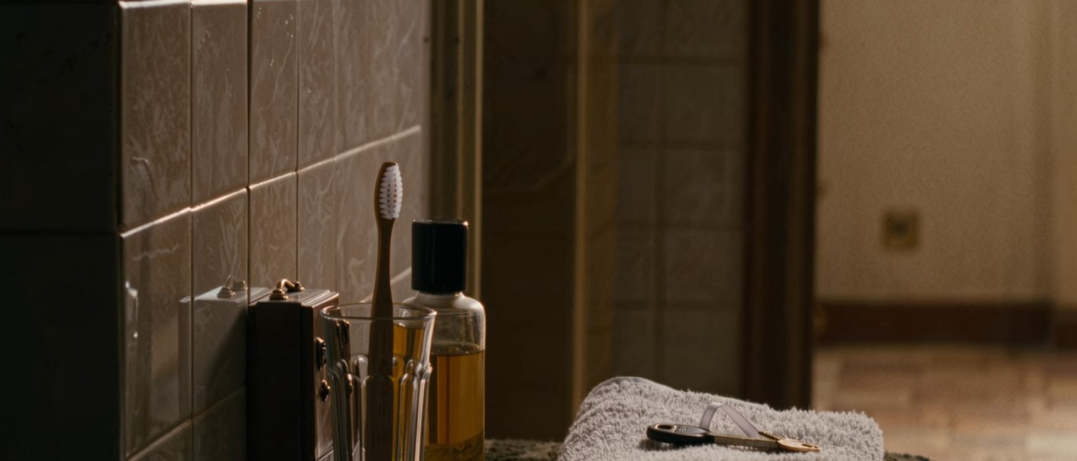 a bathroom shelf seen close with one toothbrush in a glass, a small locked box beside a bottle of scent, a folded towel and a key on its ribbon, the tiled wall close behind