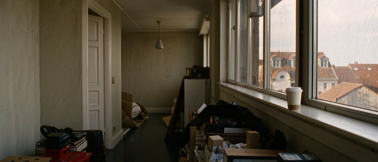 a window seat in a borrowed flat with unfamiliar rooftops beyond the glass, a coffee cup on the sill, the room behind still half packed