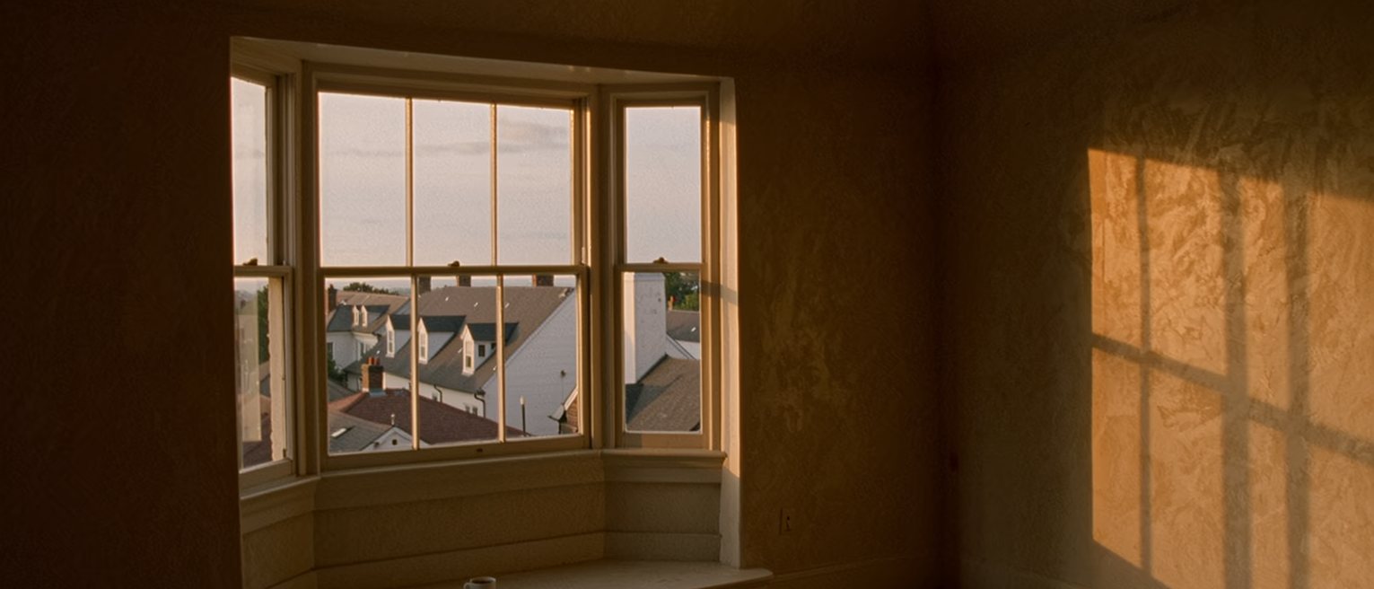 a window seat in a borrowed flat with unfamiliar rooftops beyond the glass, a coffee cup on the sill, the room behind still half packed
