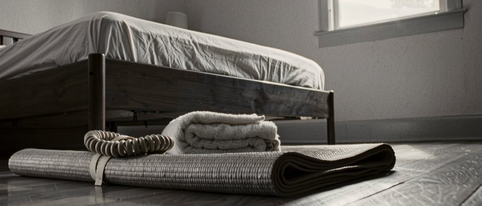 a bedroom floor seen close with a mat rolled half open, a strap coiled on it and a towel folded beside, the edge of the bed close behind