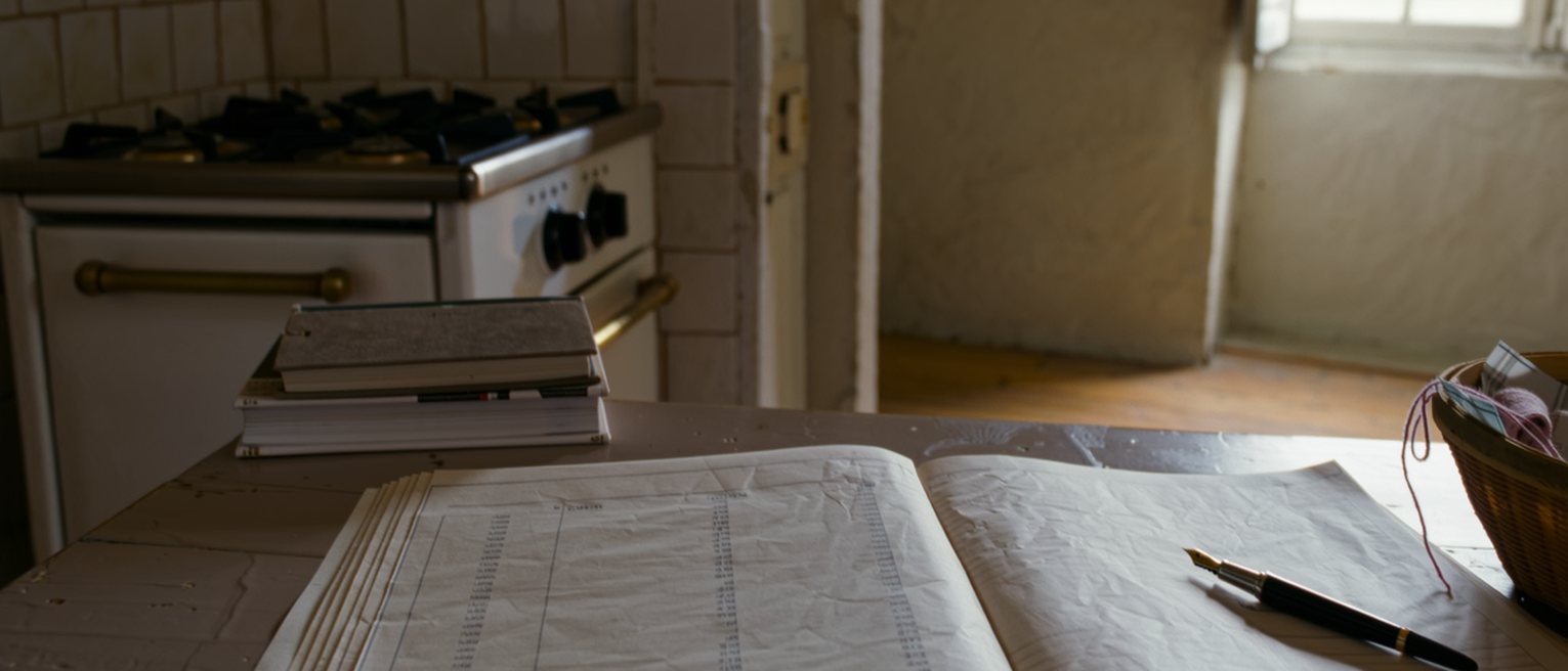 a kitchen table in a Normandy house seen close with an account ledger open beside a lined exercise book and a fountain pen, a sewing basket pushed to the far edge, a stack of new books with their spines uncracked, the stove and the tiled wall close behind and the window soft beyond