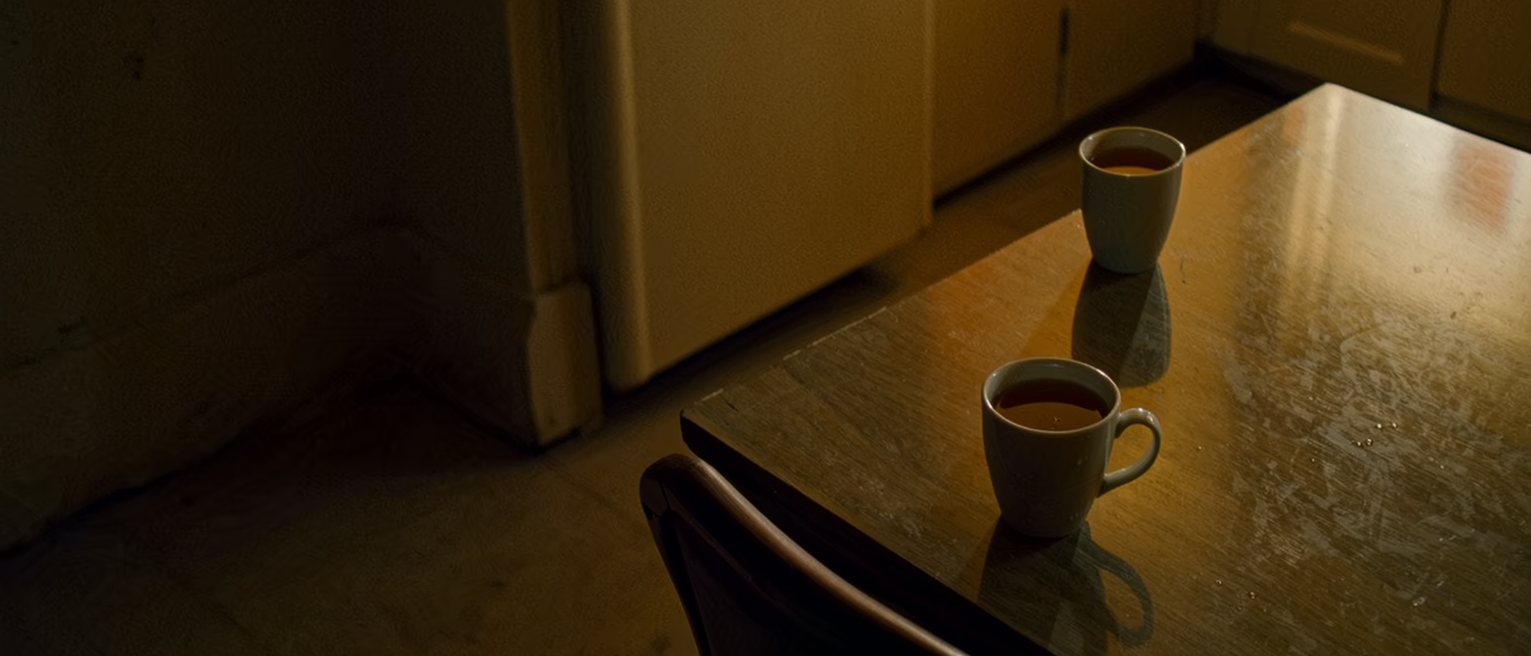 a kitchen table with one cup of tea untouched and no longer steaming, the chair before it pulled in close, and the second cup across the table still full