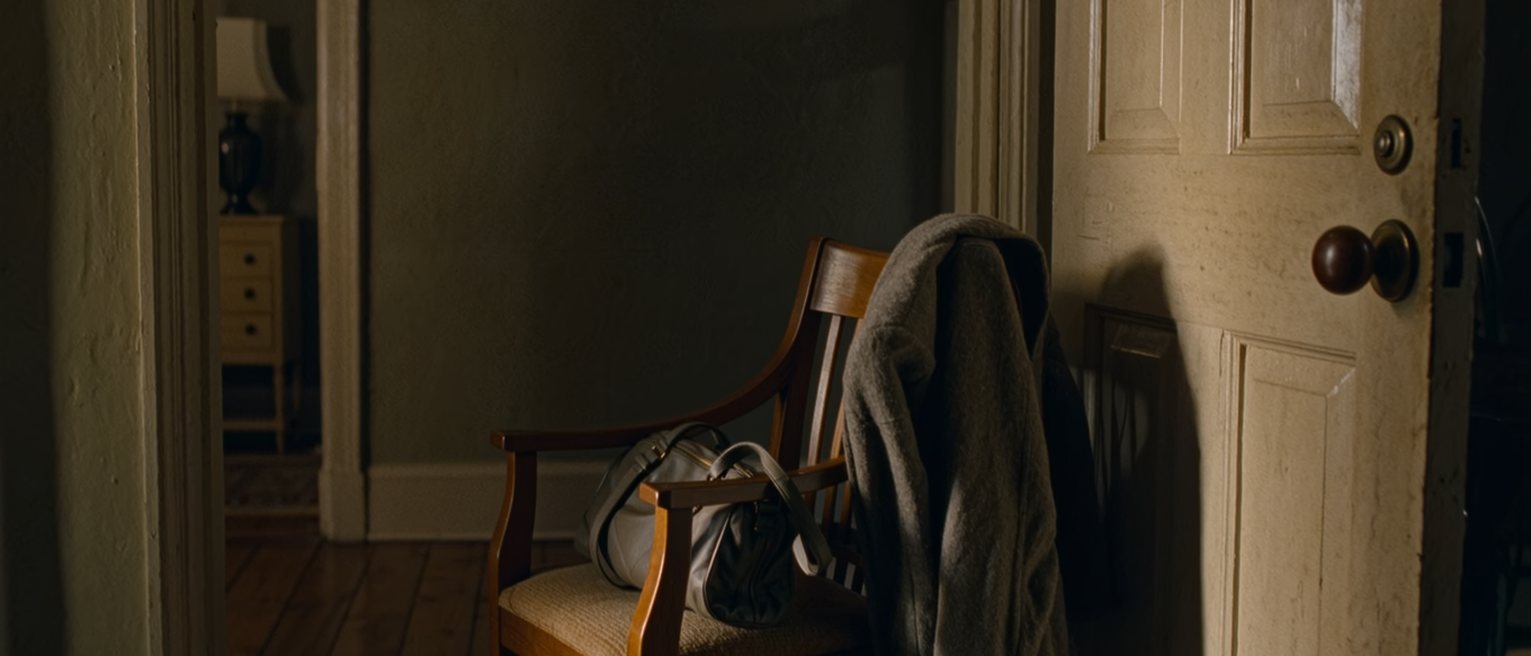 a hall chair by the front door with a coat laid over its back and a bag not yet set down, its strap still over the chair's arm