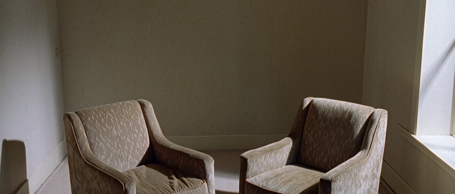 a sitting room seen close with two armchairs turned to face each other across a low table that holds one cup of tea, a small clock and a closed notebook, the cushion of the far chair still dented, a tall window soft beyond