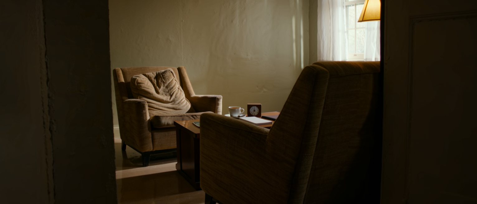 a sitting room seen close with two armchairs turned to face each other across a low table that holds one cup of tea, a small clock and a closed notebook, the cushion of the far chair still dented, a tall window soft beyond