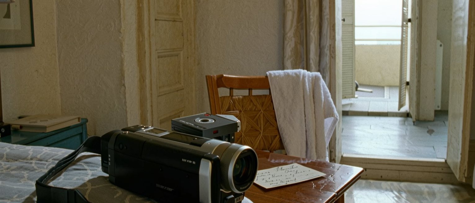 a holiday room seen close with a camcorder on the bed beside a mini-DV tape, a wet towel over the chair and a postcard half written on the table, the balcony shutters soft behind
