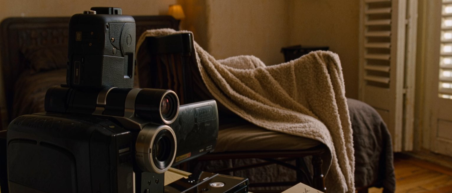 a holiday room seen close with a camcorder on the bed beside a mini-DV tape, a wet towel over the chair and a postcard half written on the table, the balcony shutters soft behind