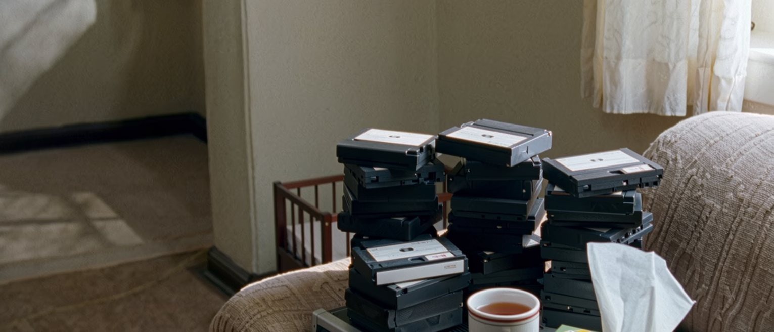 a sitting room seen close with a stack of labelled mini-DV tapes beside a cassette deck, a mug of tea and a box of tissues on the arm of the sofa, a cot at the edge and the curtain soft behind