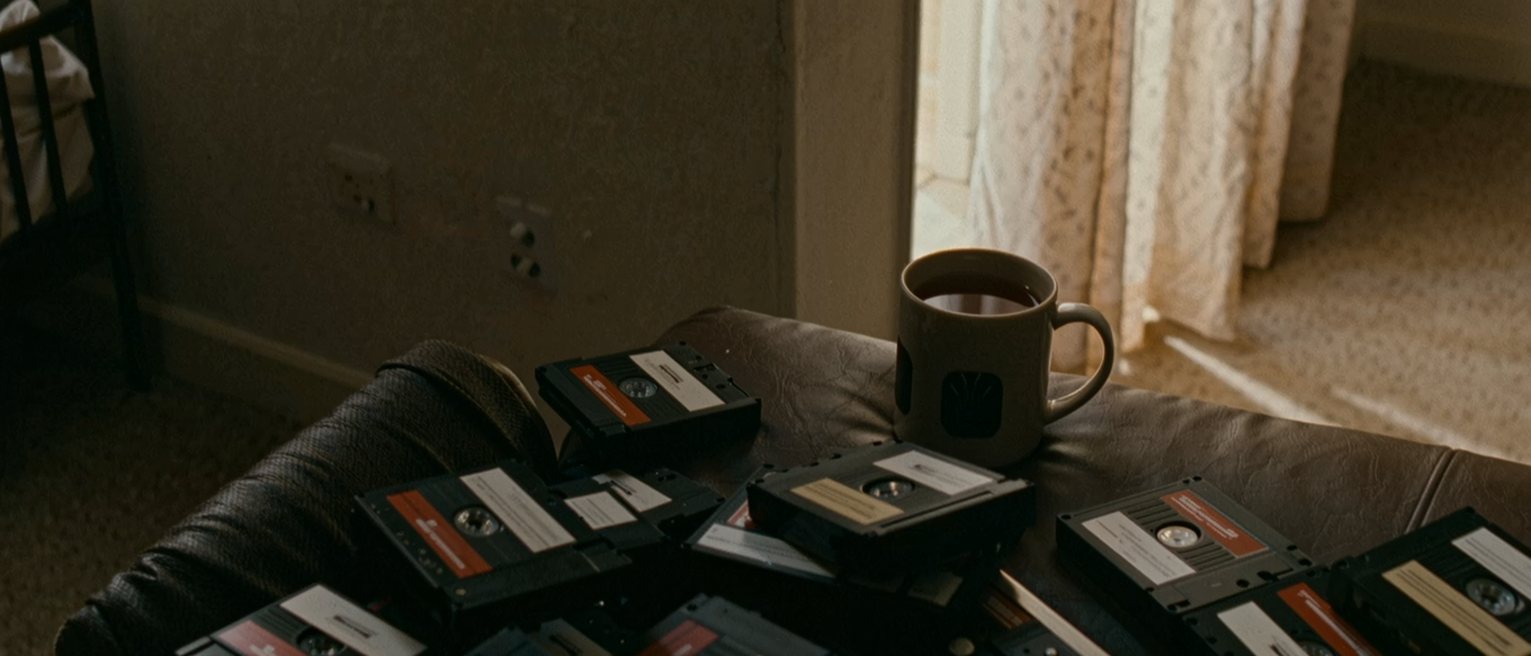 a sitting room seen close with a stack of labelled mini-DV tapes beside a cassette deck, a mug of tea and a box of tissues on the arm of the sofa, a cot at the edge and the curtain soft behind