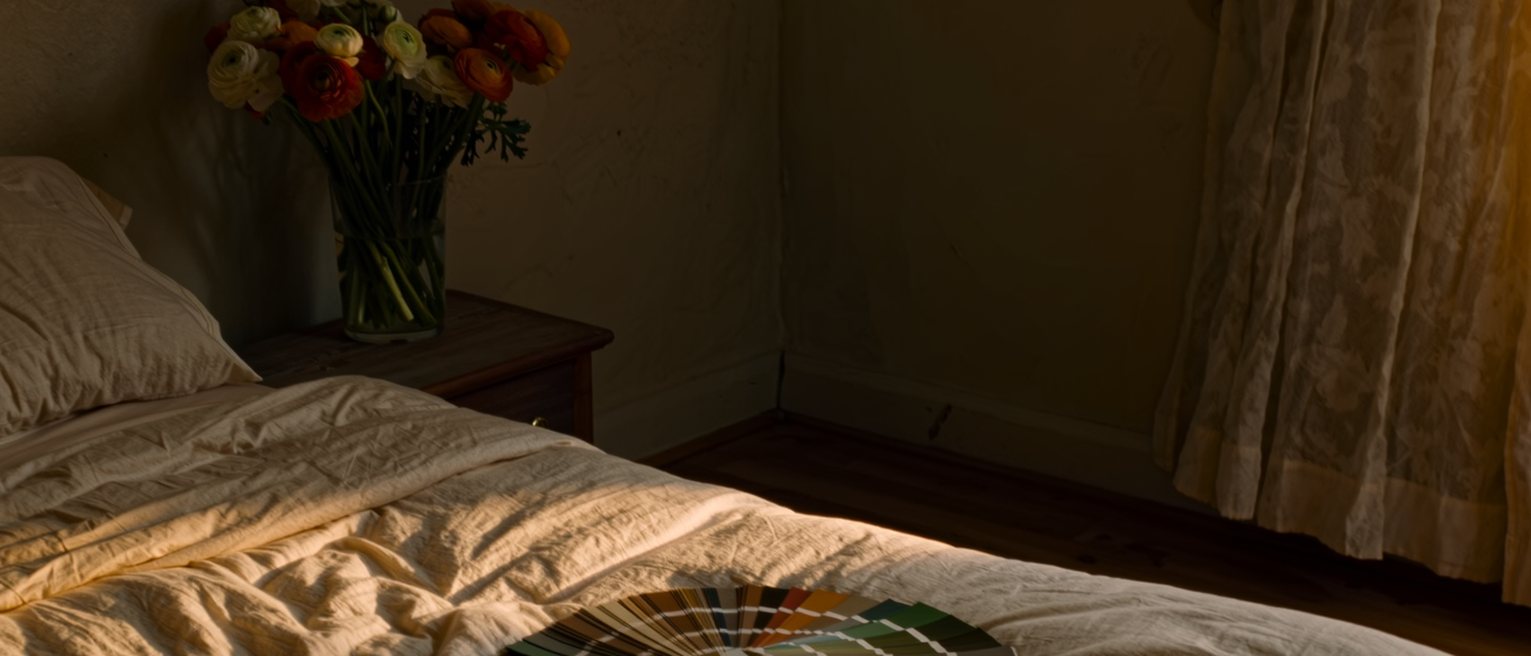a rented room seen close with the bed remade in linen sheets far too good for it, a vase of ranunculus on the bedside table, paint charts fanned on the bedspread and a car key beside them, the drawn curtain soft behind