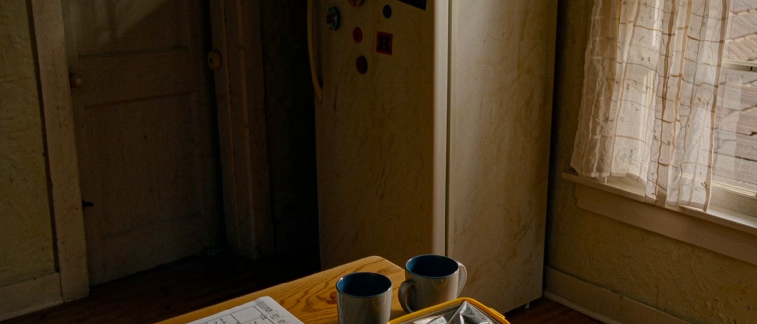 a kitchen table seen close with a sheet of terms written in two inks and a calendar with alternate weeks circled, two mugs and a packed lunchbox, the fridge with its magnets close behind and the window soft beyond