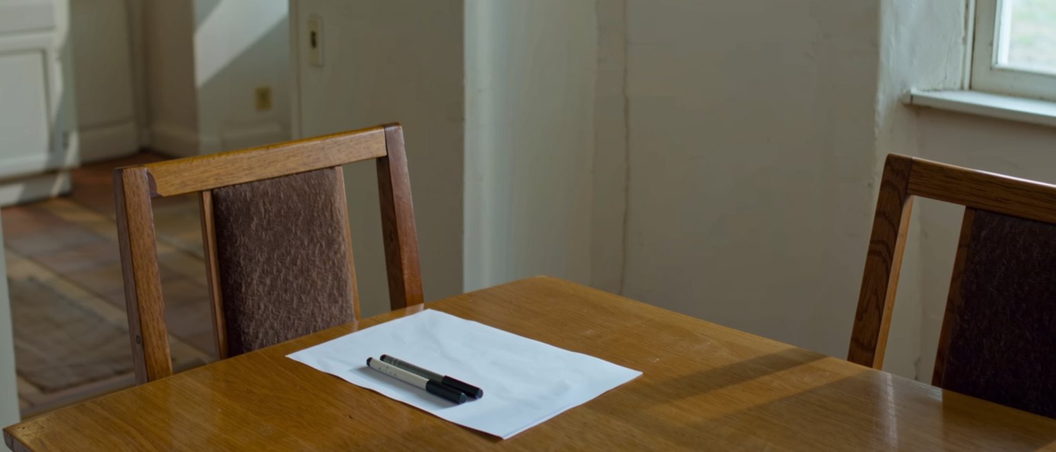 a kitchen table with a single sheet of paper set between two chairs and two pens laid down on it