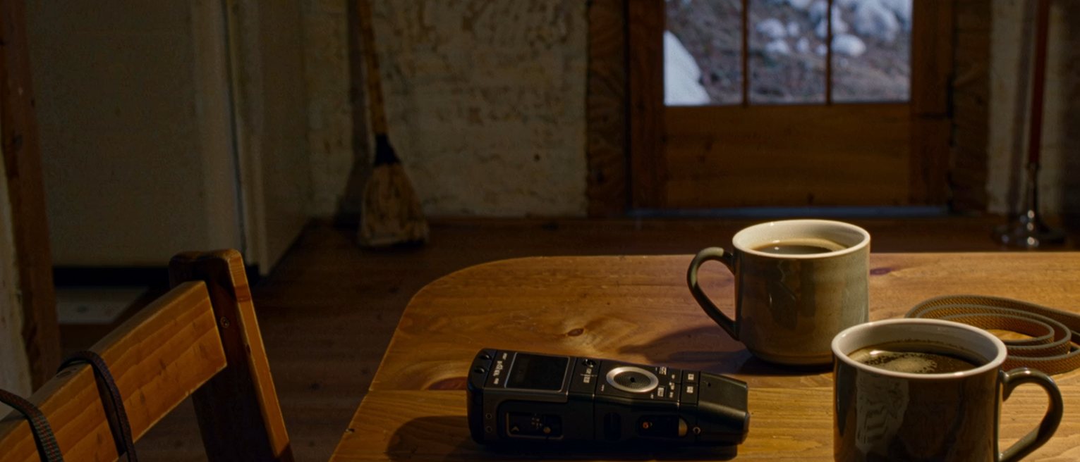 a chalet kitchen table seen close with a voice recorder beside a transcript bound with a clip, two cups of coffee and a dog lead coiled on the chair, the snow on the glass soft behind