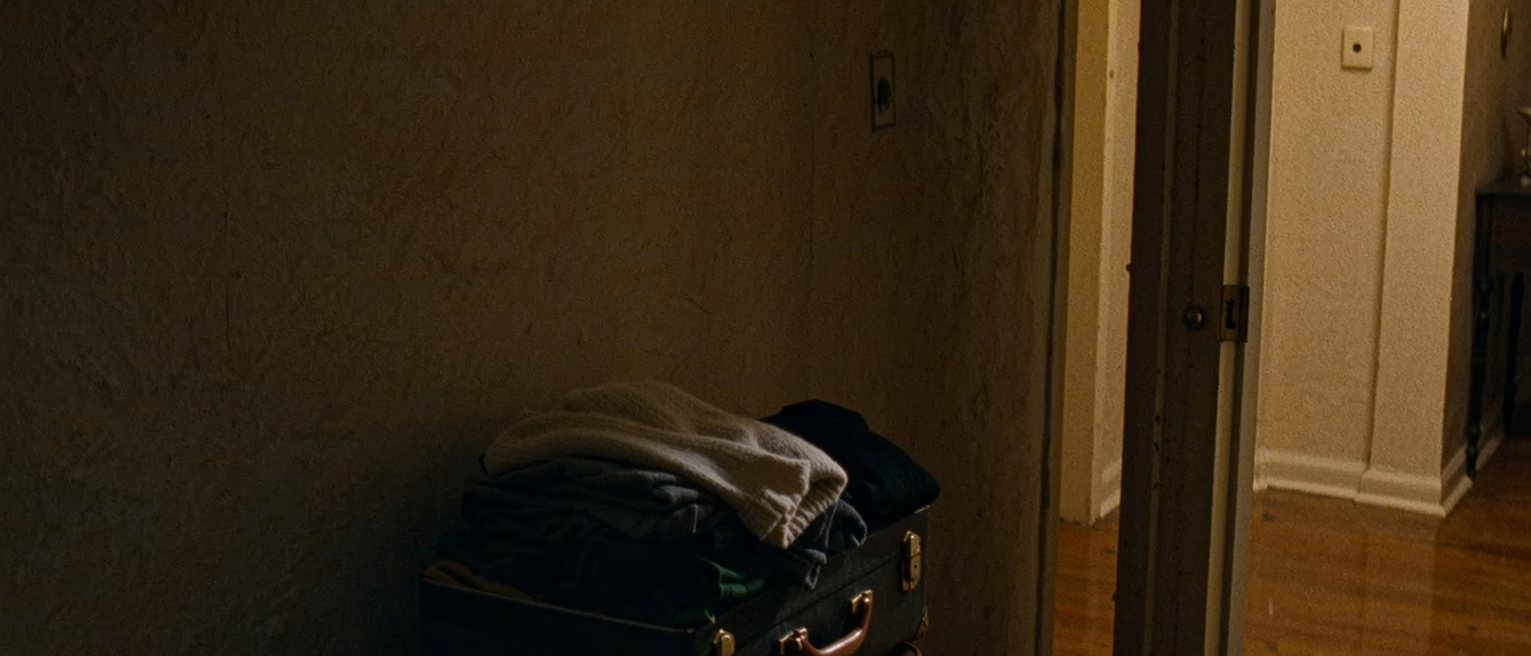 a hall seen close, a suitcase packed and set by the door standing open onto a corridor, a set of keys and a folded coat on the hall table beside it
