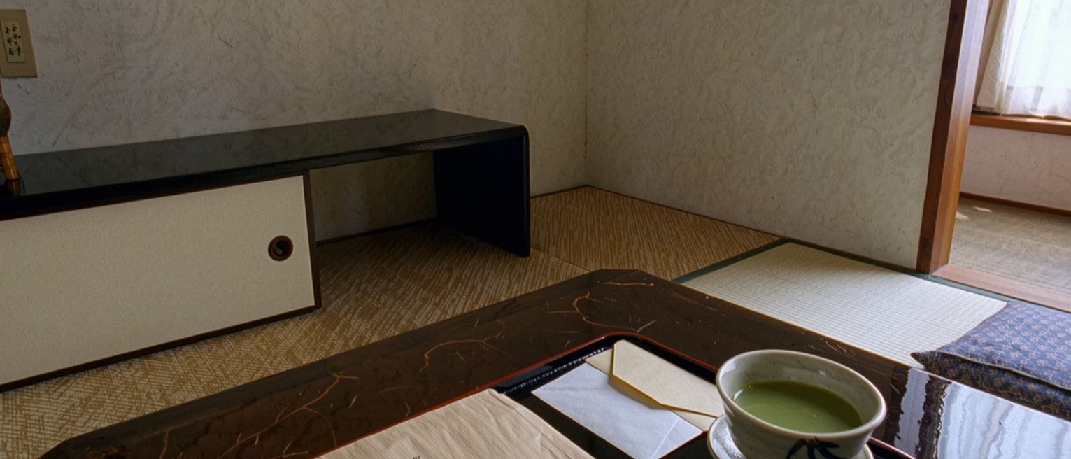 a low table seen close with a letter in calligraphy lying open beside its envelope and a red seal, a folded furoshiki and a small box of wagashi, the tatami and the tokonoma with its single vase close behind