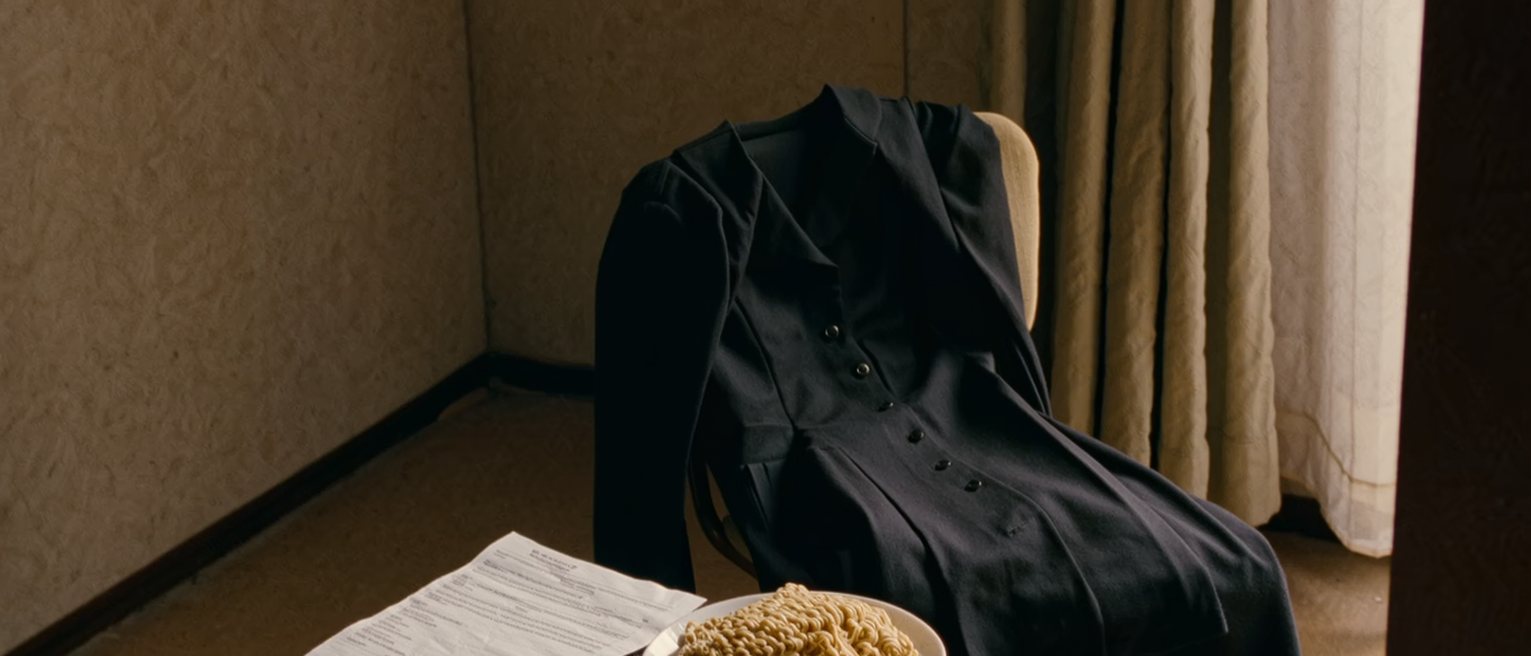 a one-room flat seen close with a tuition statement on the small table, a bowl of instant noodles, a bus ticket home to Toyama and a folded black dress over the chair, the curtain soft behind