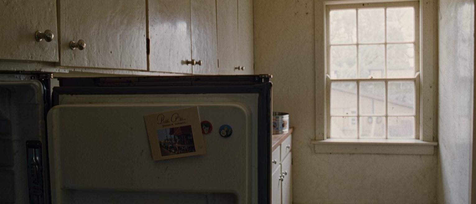 a kitchen seen close with a fridge door crowded with an invitation, a pinned postcard and two magnets, a cake tin on the counter and the window soft behind