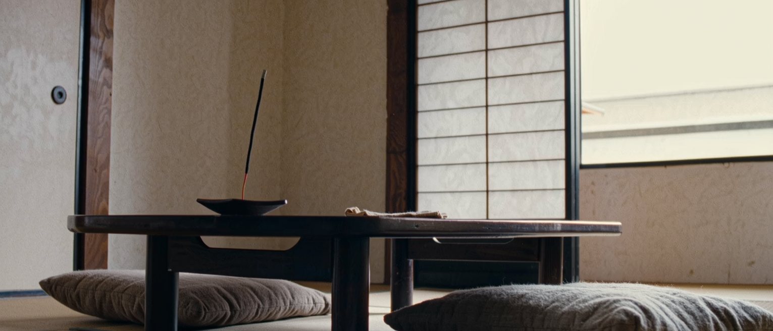 a low table seen close with two cushions set either side of it, a single stick of incense burned halfway down in its holder and a folded cloth, the shoji screen soft behind