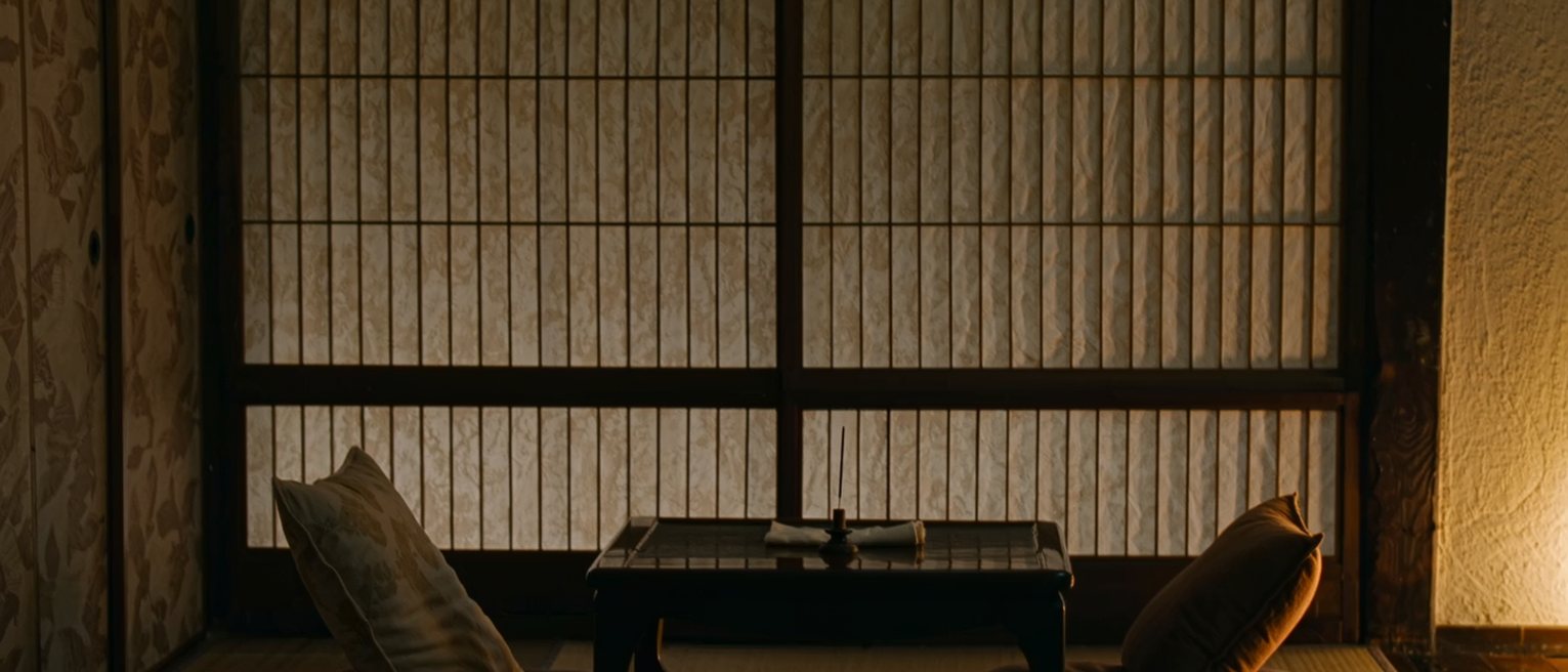 a low table seen close with two cushions set either side of it, a single stick of incense burned halfway down in its holder and a folded cloth, the shoji screen soft behind