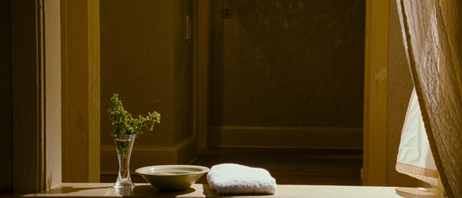 a windowsill seen close with a bowl of water and a folded white towel beside it, a sprig of green in a glass, the curtain moving a little in the draught behind