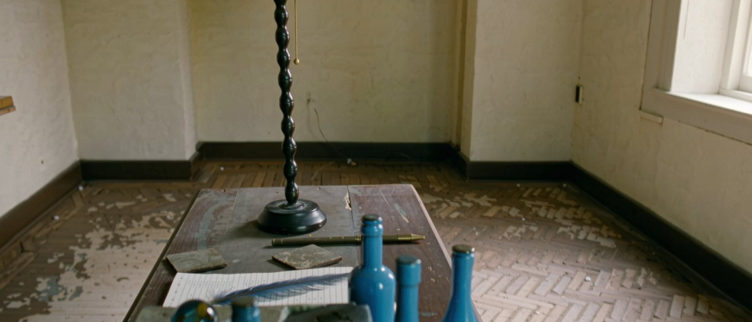 a desk seen close with a row of blue things laid out, a bottle, a glass marble, a shard of tile and a feather, beside a notebook with numbered lines, the lamp close behind