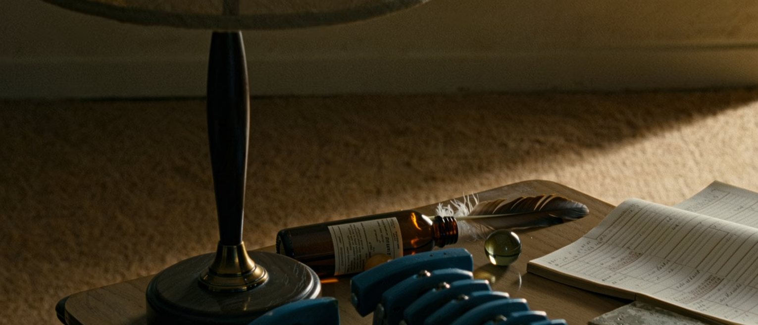 a desk seen close with a row of blue things laid out, a bottle, a glass marble, a shard of tile and a feather, beside a notebook with numbered lines, the lamp close behind