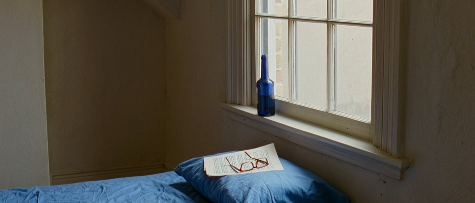 a bedroom seen close with a blue bedspread and a blue bottle on the sill, a numbered manuscript on the pillow and a pair of reading glasses, the window close behind