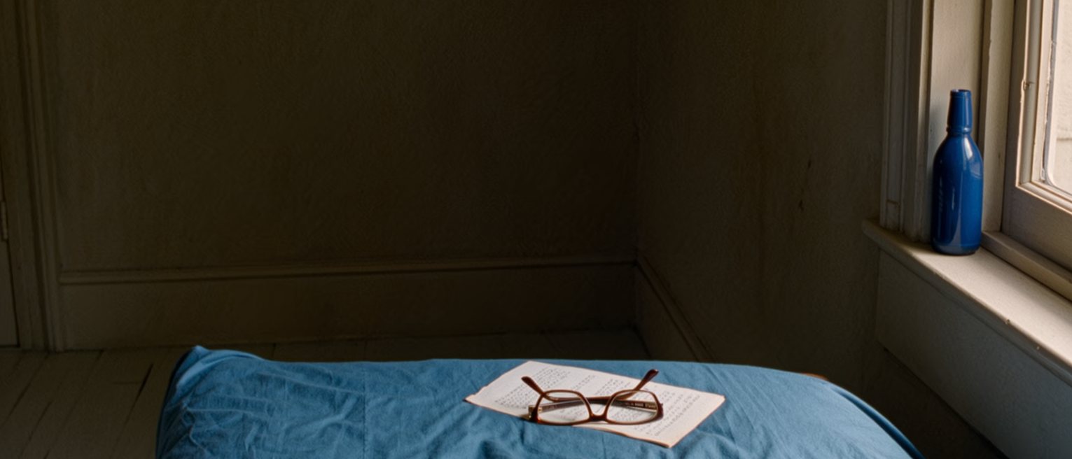 a bedroom seen close with a blue bedspread and a blue bottle on the sill, a numbered manuscript on the pillow and a pair of reading glasses, the window close behind