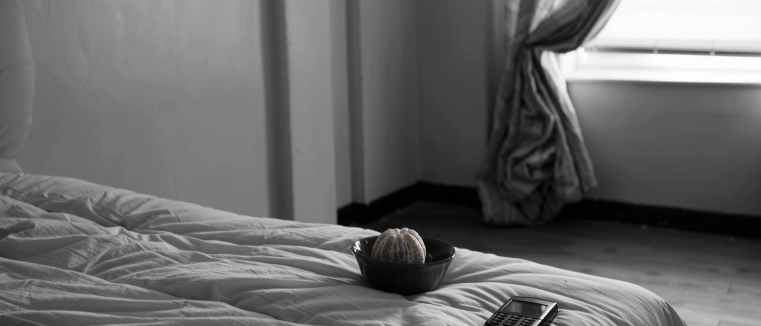 a bedroom with the curtains drawn and a phone switched off on the far side of the room, a bowl with a peeled orange set on the bed