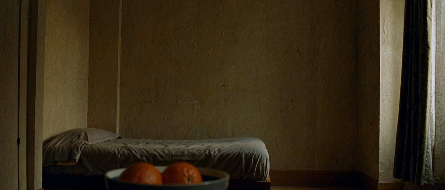 a bedroom with the curtains drawn and a phone switched off on the far side of the room, a bowl with a peeled orange set on the bed