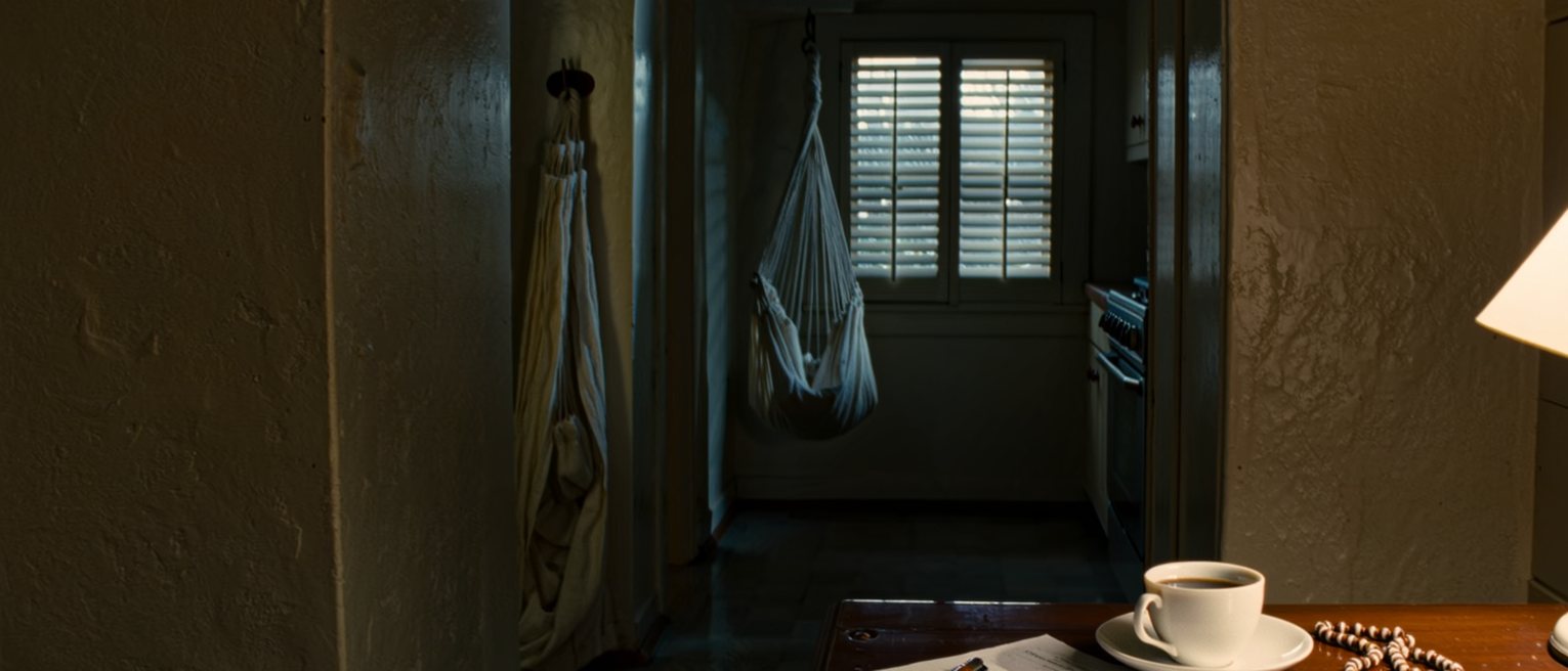 a kitchen table seen close with a census form filled in pen, a rosary and a cup of coffee, a hammock hook on the wall and the window shutters soft behind