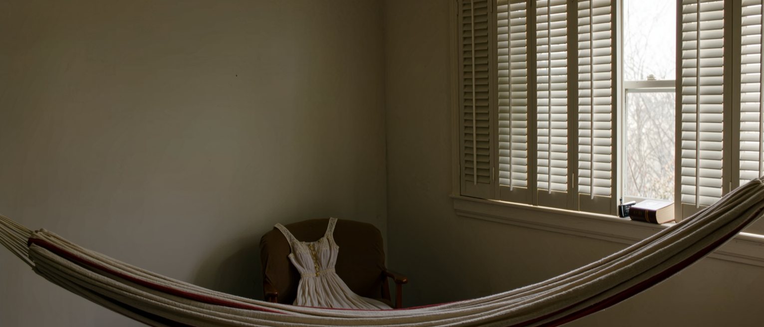 a bedroom seen close with a hammock slung across the corner, a dress on the chair and a bible and a phone side by side on the sill, the shutters open behind
