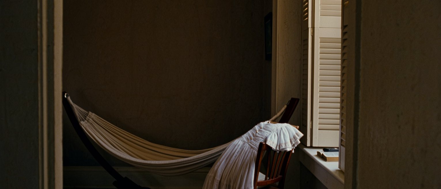 a bedroom seen close with a hammock slung across the corner, a dress on the chair and a bible and a phone side by side on the sill, the shutters open behind