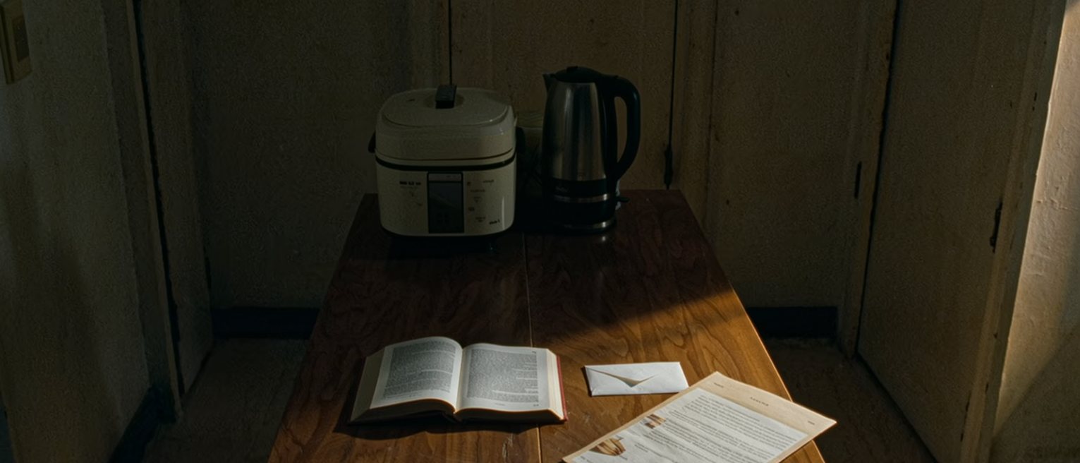 a kitchen table seen close in a small flat with a paperback open face down, a pay envelope and a leaflet from a clinic beside it, a rice cooker and a kettle close behind