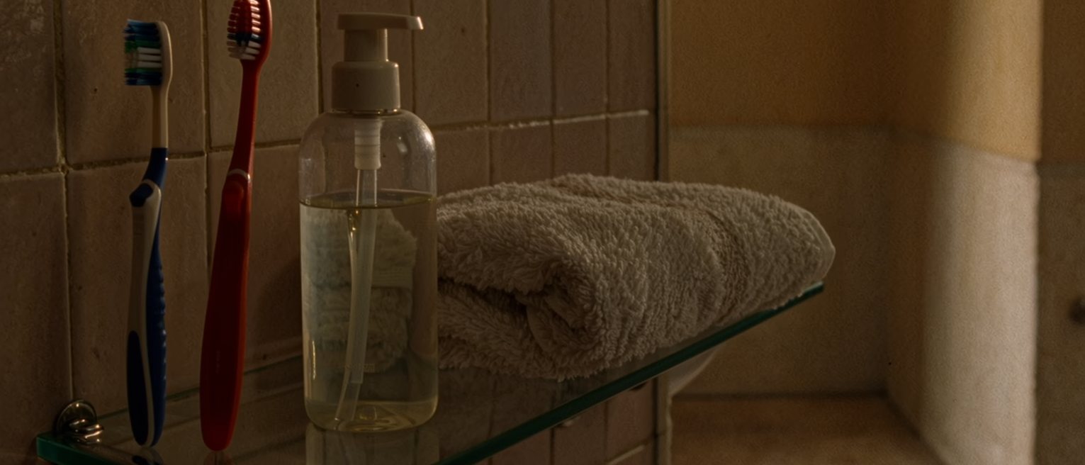 a bathroom shelf seen close with a child's toothbrush and a grown one in the same glass, a bottle of shampoo and a folded towel, the tiled wall soft behind