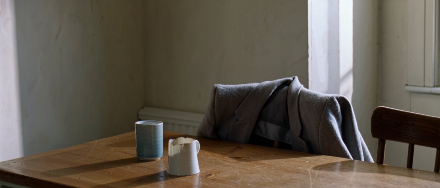 a kitchen table at home with two cups on it, one drunk to the bottom and one untouched, and a coat still hung over the back of the second chair