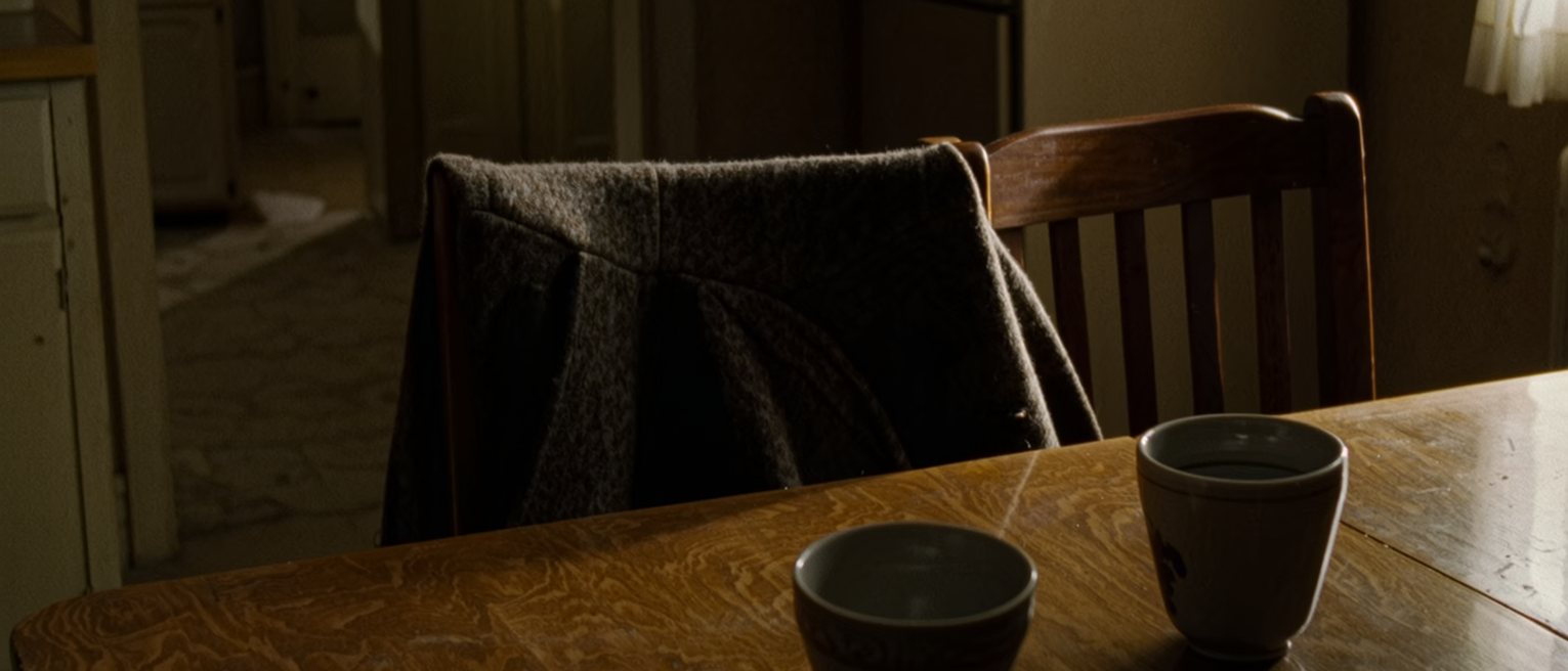 a kitchen table at home with two cups on it, one drunk to the bottom and one untouched, and a coat still hung over the back of the second chair