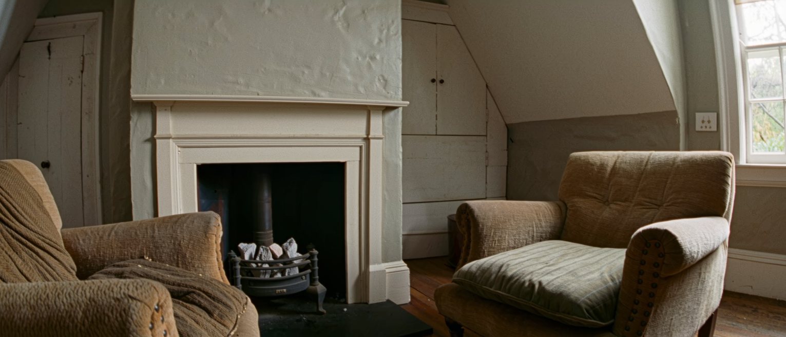 two armchairs drawn up close to a tidy domestic grate where the coals have burned low, one cushion still holding a deep indent and the other unmarked