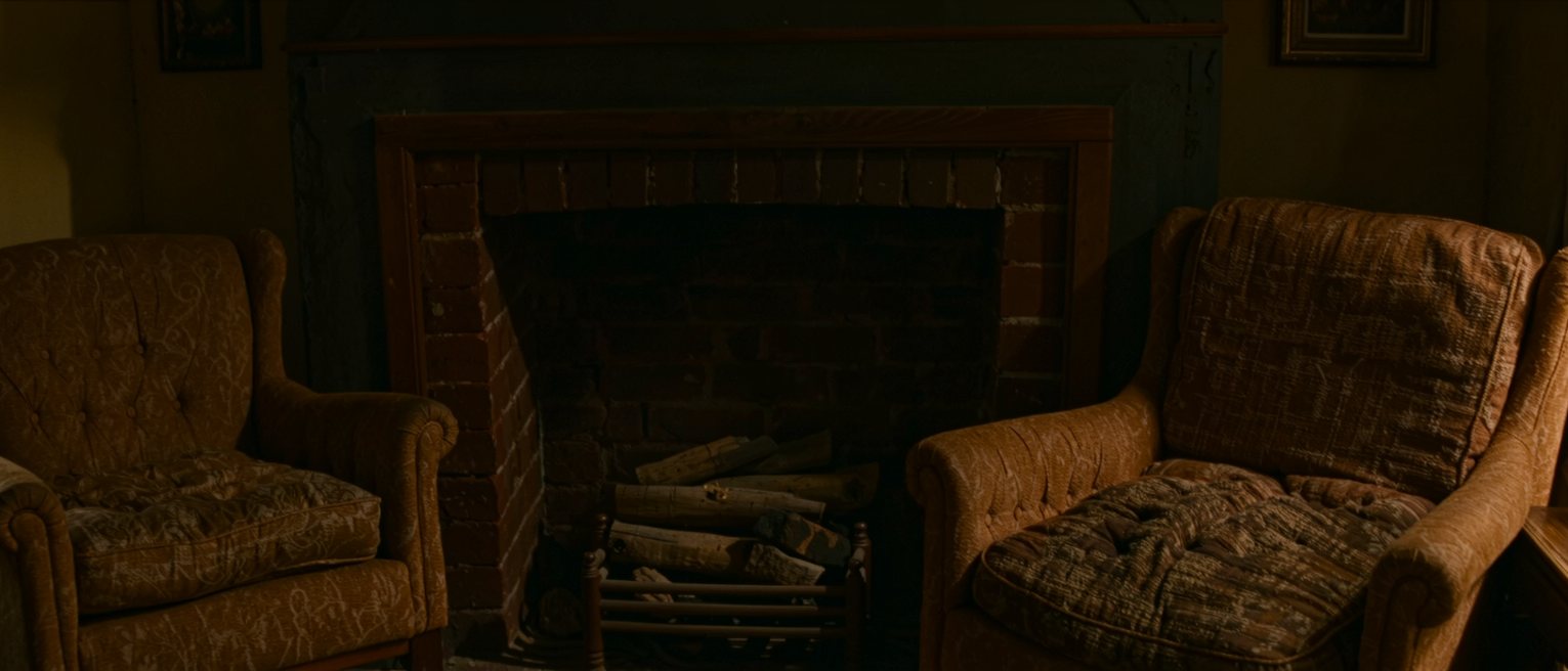 two armchairs drawn up close to a tidy domestic grate where the coals have burned low, one cushion still holding a deep indent and the other unmarked