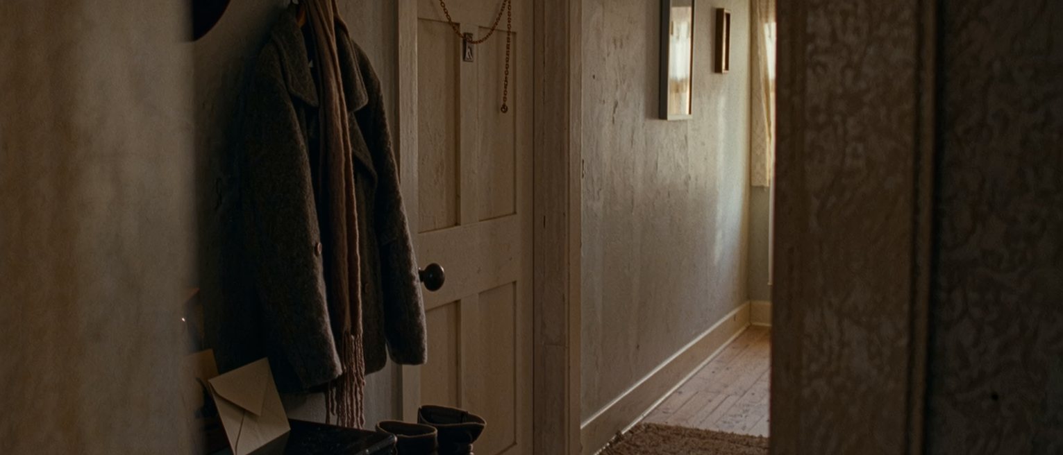 a hall seen close with a winter coat and a long scarf on the hooks, a pair of boots on the mat and an envelope propped on the console, the door with its chain soft behind