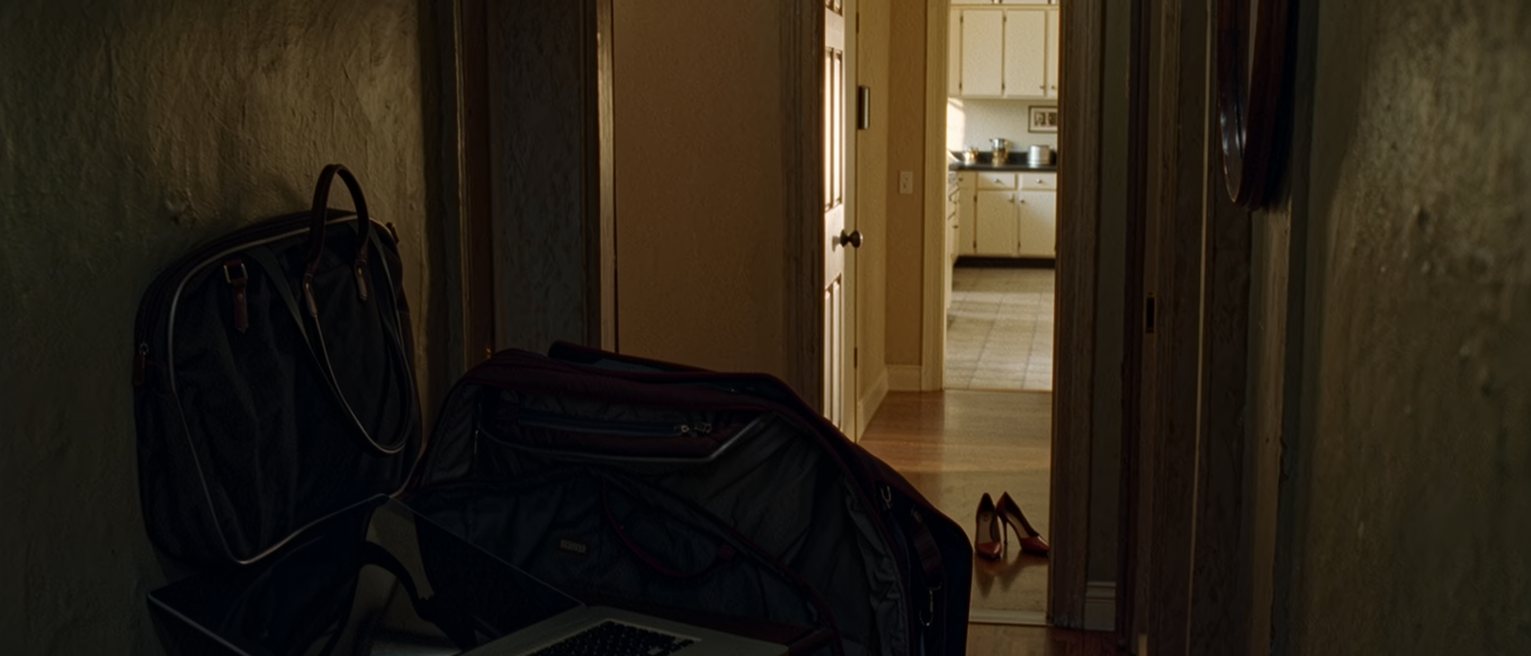 a hall seen close with a work bag dropped by the door still open with its laptop shut inside, a set of keys on the console and a pair of heels stepped out of, the lit kitchen doorway soft behind